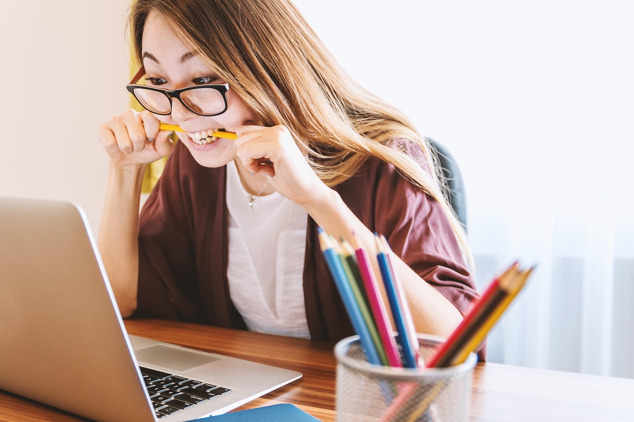 woman biting pencil while looking at laptop